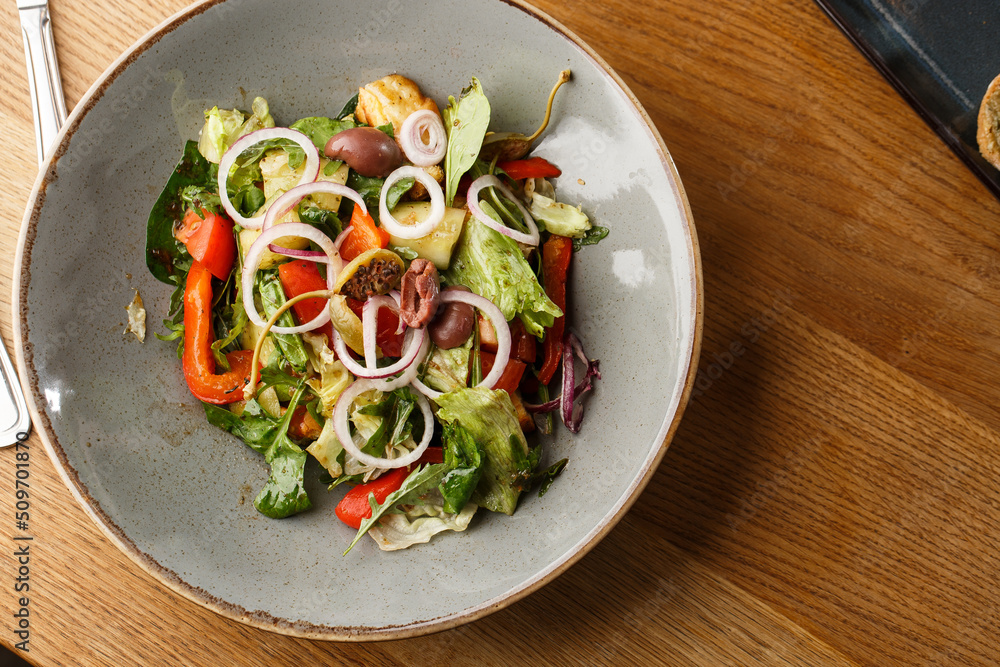 Greek salad with fresh vegetables: tomato, cucumber, red bel pepper, lettuce, onion, olives and cheese. Close-up on a white round plate on a wooden background. Salad menu with ingredients