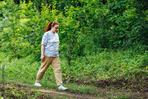 Obraz na plátně Side view of young plump buxom woman with long curly red hair in ponytail, wearing blue blouse with floral pattern, beige trousers, walking on path in park forest among green trees