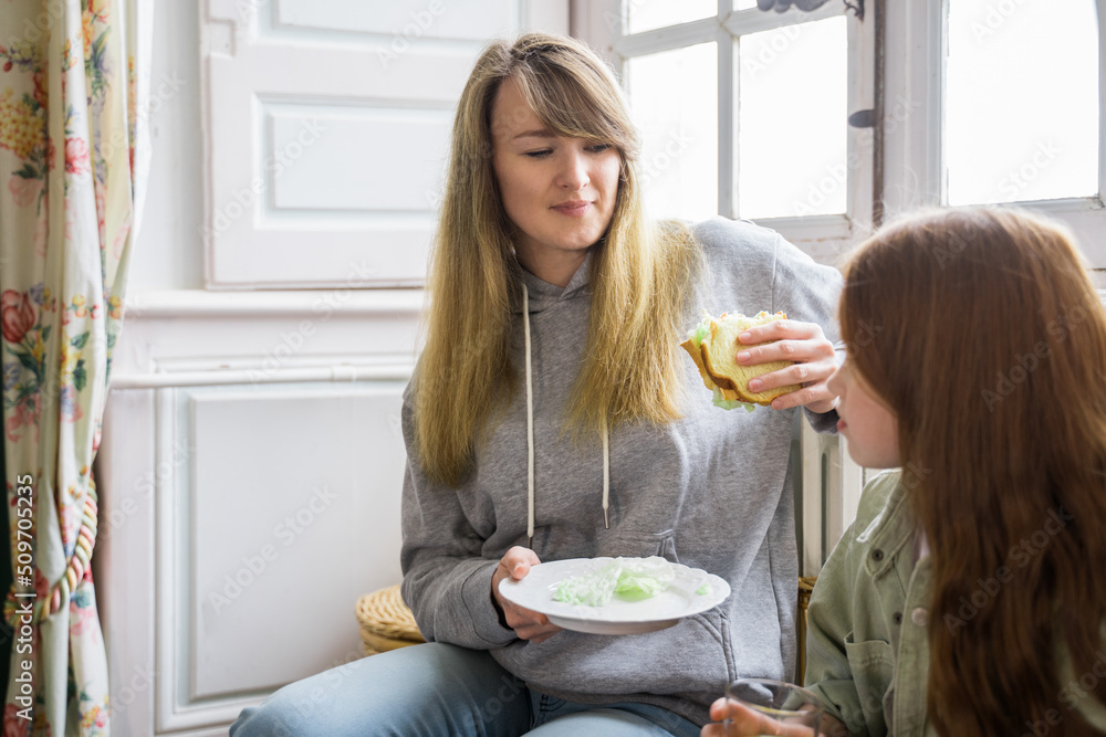Caucasian woman holding sandwich and looking at her daughter while sitting at the room Caucasian woman holding sandwich and looking at her daughter while sitting at the room