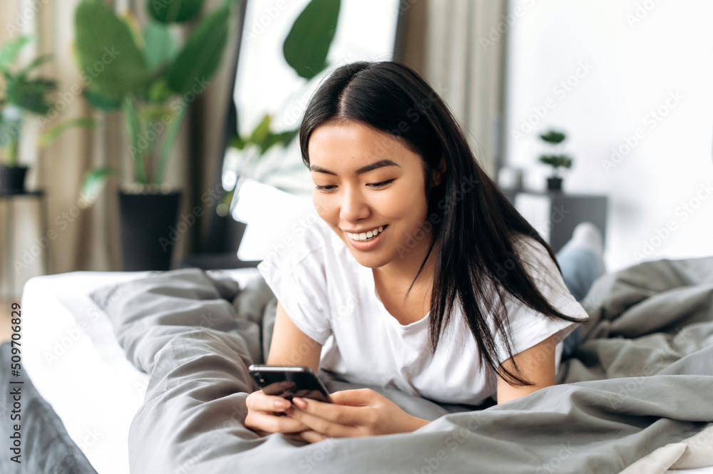 Pretty happy asian brunette girl dressed in a basic white t-shirt, is laying on a bed in a room, uses her cell phone, chatting with friends in social media, checking email, browsing news, smiling Pretty happy asian brunette girl dressed in a basic white t-shirt, is laying on a bed in a room, uses her cell phone, chatting with friends in social media, checking email, browsing news, smiling