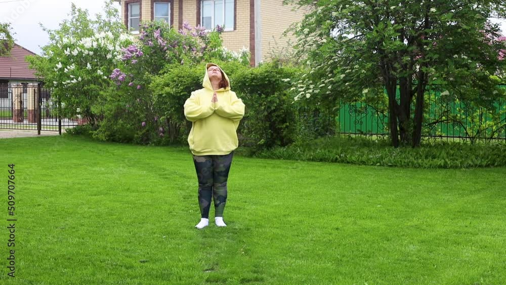 Fat and overweight woman in yellow hoodie doing breathing exercises, fitness with hands together, turning head left and right in the green backyard outdoors. Nature, park and house on background. Fat and overweight woman in yellow hoodie doing breathing exercises, fitness with hands together, turning head left and right in the green backyard outdoors. Nature, park and house on background.