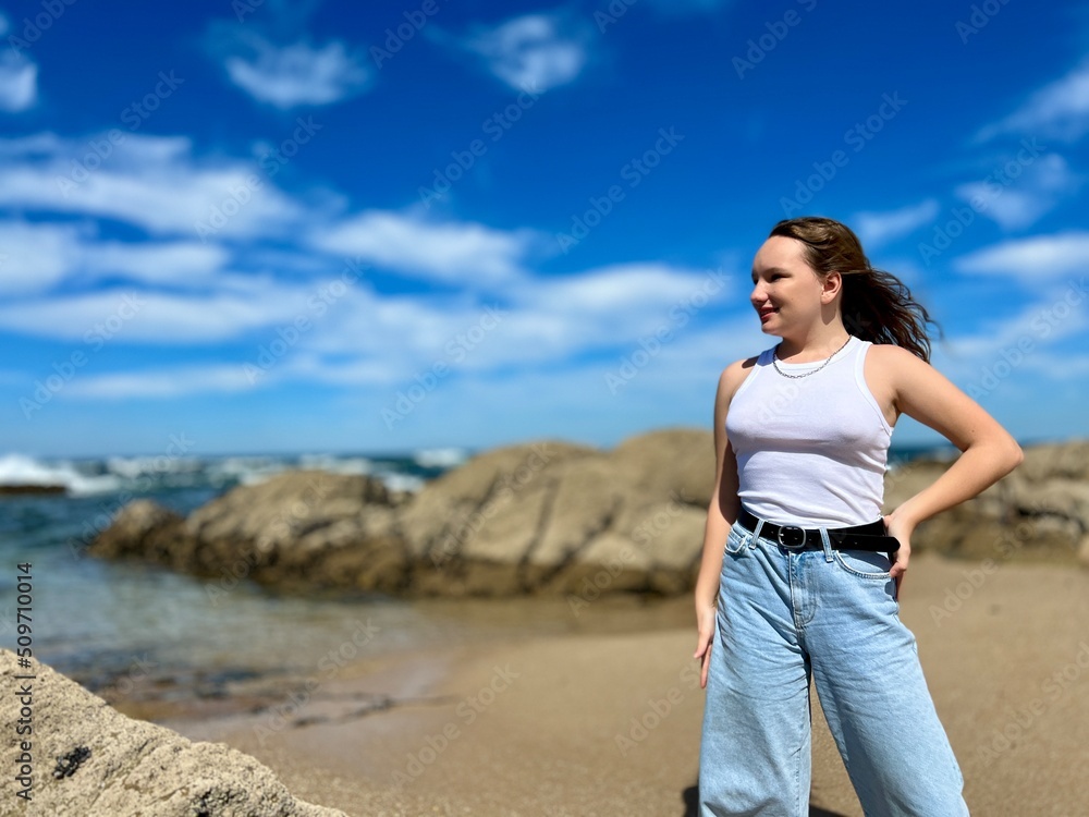 a girl in blue jeans and a white T-shirt stands in Portugal against the backdrop of the Atlantic Ocean city of Panta de Lima in the background stones and waves of the ocean There is a place for text.