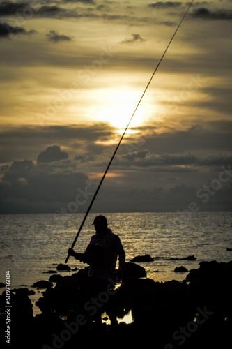 Scenic View Of Portraits Man Silhouette Walking With A Fishing Rod In The Twilight Sun On A Rocky Beach