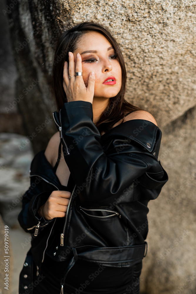 Confident woman  with red lips  in trendy black leather jacket and sensual black dress posing on rocky background.