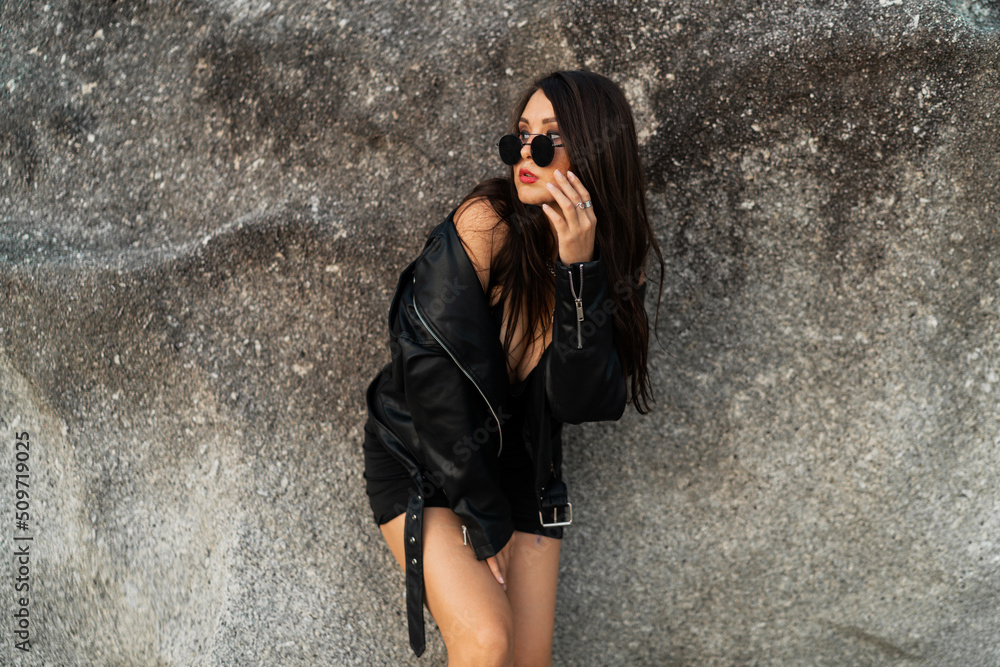 Gorgeous asian woman with red lips in trendy black leather jacket and sensual black dress posing on rocky background. Gorgeous asian woman with red lips in trendy black leather jacket and sensual black dress posing on rocky background.