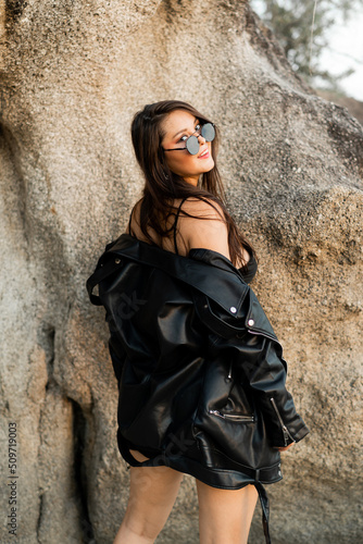 Fashionable sensual woman in black leather jacket and stylish sunglasses posing over rocks.