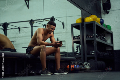 Young African American male looking at his phone to reply to an online message. Happy, mixed race male smiling at his phone while sitting down in the gym. High quality photo 