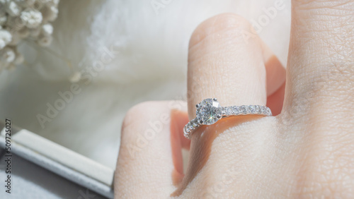 Close up of diamond ring on woman’s finger with  white flower, sunlight and shadow background. Love, valentine, relationship and wedding concept. Soft and selective focus.