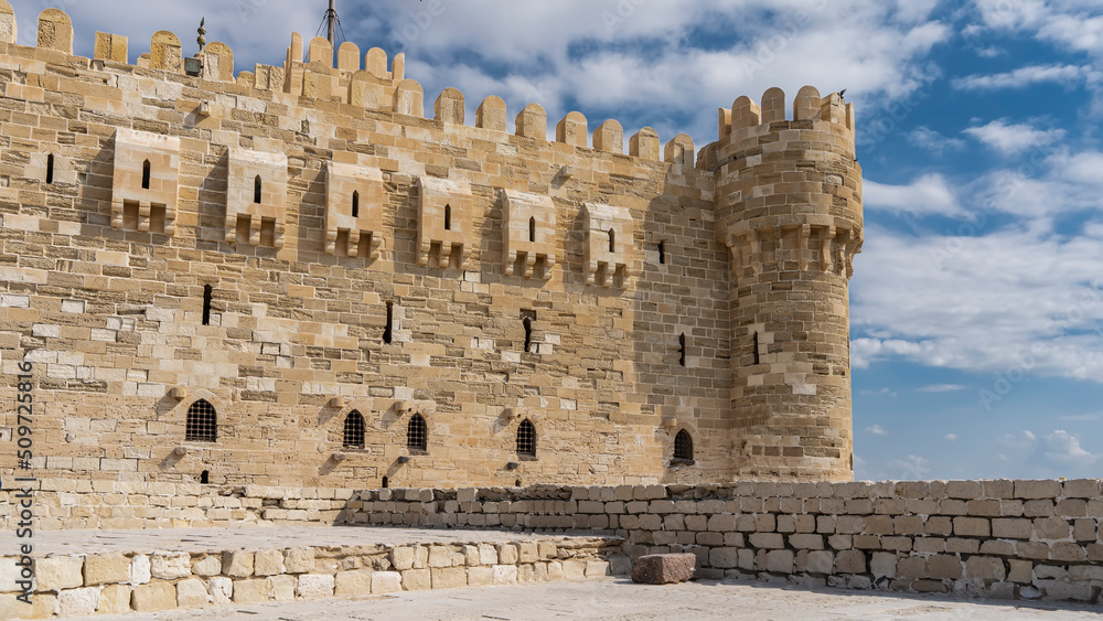 A fragment of the ancient Citadel of Qaitbay. Visible masonry walls, corner tower, loopholes, barred windows. Blue sky with clouds. Egypt. Alexandria