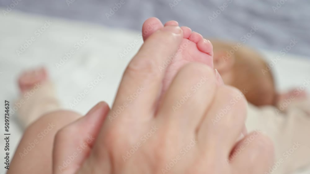 Mother caresses bare tiny feet of newborn daughter lying on bed on blurred background. Hands of woman touching soft skin of baby girl extreme closeup Mother caresses bare tiny feet of newborn daughter lying on bed on blurred background. Hands of woman touching soft skin of baby girl extreme closeup