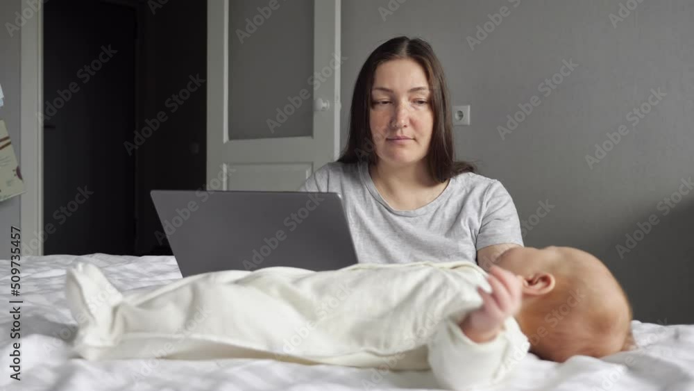 Mother pays attention to little baby girl sitting near bed and working on laptop. Newborn child tries to draw attention of mom lying on parent bed Mother pays attention to little baby girl sitting near bed and working on laptop. Newborn child tries to draw attention of mom lying on parent bed
