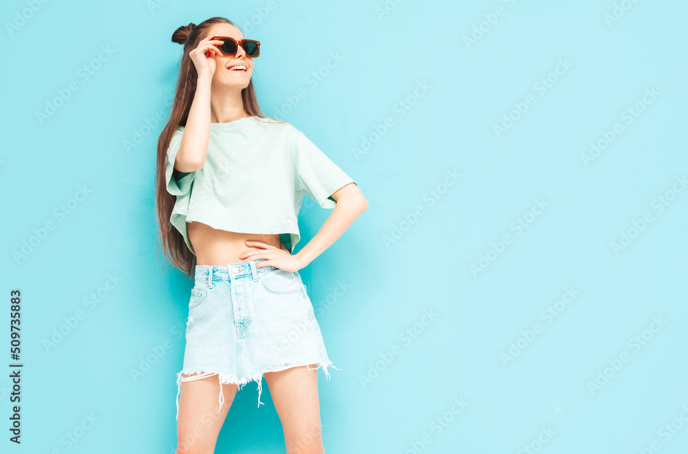 Portrait of young beautiful smiling female in trendy summer jeans skirt. carefree woman posing near blue wall in studio. Positive model having fun indoors. Cheerful and happy. in sunglasses Portrait of young beautiful smiling female in trendy summer jeans skirt. carefree woman posing near blue wall in studio. Positive model having fun indoors. Cheerful and happy. in sunglasses