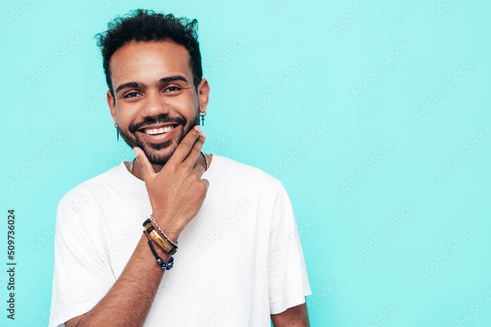Handsome smiling hipster model. Sexy unshaven man dressed in white summer t-shirt and jeans clothes. Fashion male posing near blue wall in studio. Isolated. Touching his chin Handsome smiling hipster model. Sexy unshaven man dressed in white summer t-shirt and jeans clothes. Fashion male posing near blue wall in studio. Isolated. Touching his chin