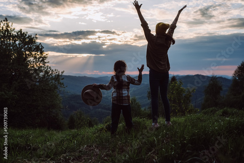 Young mother and little daughter child hikers jumping on background sunset in mountains. Family raising up hands on high rock in evening nature. Tourism, traveling and healthy lifestyle concept