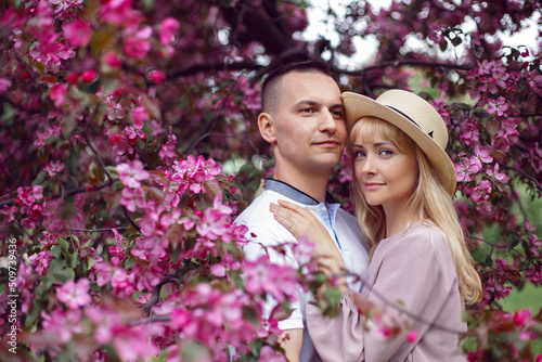 portrait man and a woman in hat stand by a blooming pink cherry tree in summer