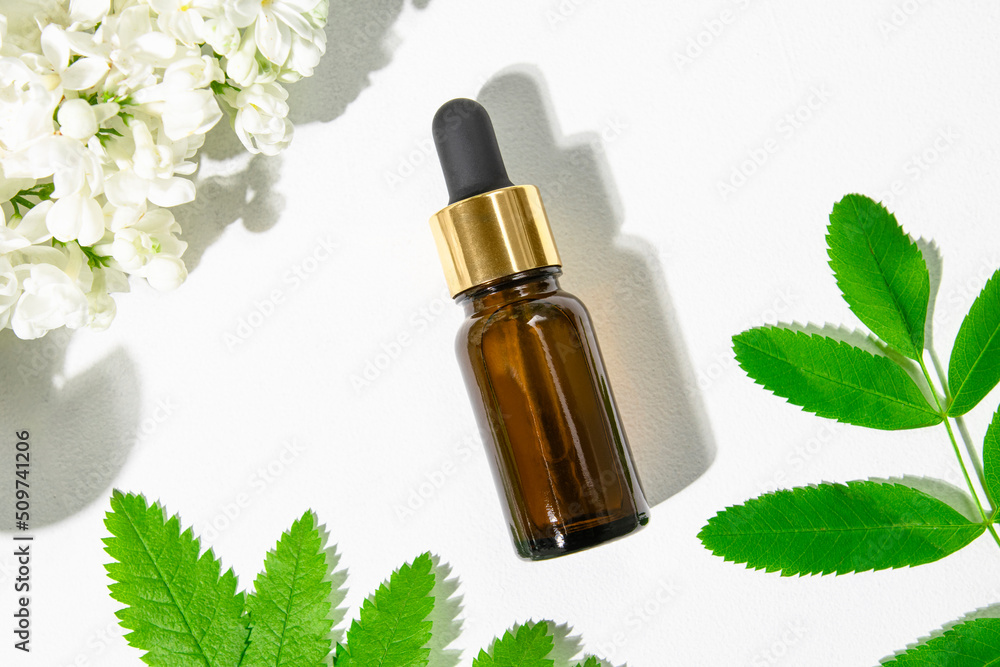 Brown glass bottle with cosmetic serum, essential oil on a white background with flowers and green leaves of the plant, top view. Aromatherapy, herbal medicine.