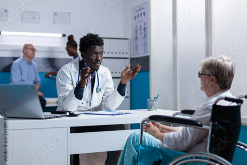 Physician expert explaining noninvasive procedure and treatment schedule to senior patient in wheelchair. Person with disability listening to hospital doctor talking about checkup appointment