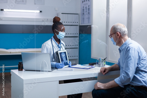 African american radiology specialist explaining x-ray scan image to senior patient while wearing virus protection facemasks. Expert radiologist showing MRI scan image to elderly man.
