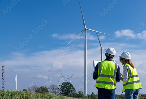 Engineer wearing uniform ,helmet inspection and survey work in wind turbine farms rotation to generate electricity energy. Green ecological power energy generation wind sustainable energy concept.