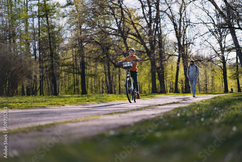 Caucasian little boy riding a bicycle with basket. Father walking in distance