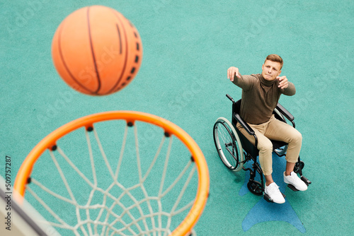 Concentrated young man with paralyzed legs sitting in wheelchair and biting lip while throwing ball in basket on sports ground
