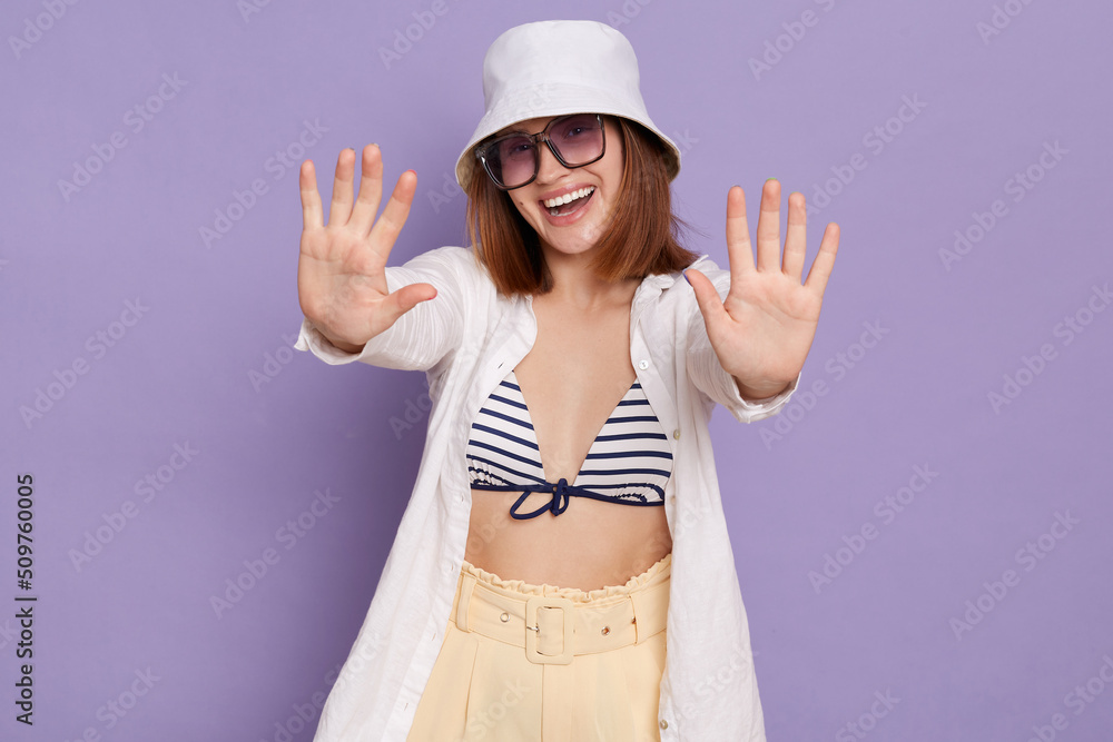 Portrait of satisfied Caucasian attractive woman wearing summer clothing and panama posing isolated over purple background, standing with raised arms, giving hive five, enjoying her rest. Portrait of satisfied Caucasian attractive woman wearing summer clothing and panama posing isolated over purple background, standing with raised arms, giving hive five, enjoying her rest.
