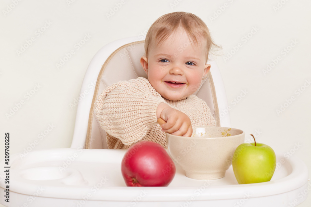 Horizontal shot of smiling charming toddler baby girl dresses in beige jumper sitting in high chair and eating fruit or vegetable puree, looking at camera, isolated over white background Horizontal shot of smiling charming toddler baby girl dresses in beige jumper sitting in high chair and eating fruit or vegetable puree, looking at camera, isolated over white background