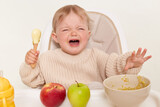 Indoor shot of sad upset infant baby girl dresses in beige jumper sitting in high chair and and crying, doesn't want to eat porridge, isolated over white background