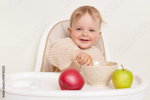 Horizontal shot of smiling charming toddler baby girl dresses in beige jumper sitting in high chair and eating fruit or vegetable puree, looking at camera, isolated over white background