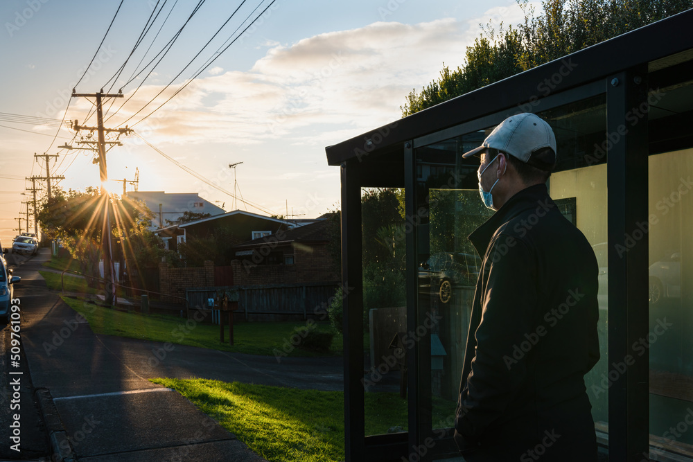 Man wearing a face mask waiting for bus at a suburb bus stop with morning sunbursts shining through the street power lines Man wearing a face mask waiting for bus at a suburb bus stop with morning sunbursts shining through the street power lines
