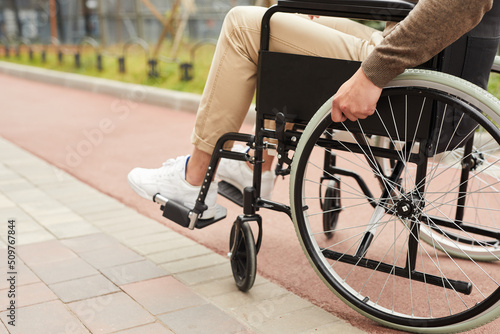 Close-up of unrecognizable man in beige jeans having difficult to walk sitting in modern wheelchair outdoors