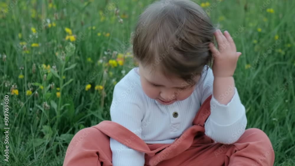 Close-up portrait of cute little girl child sitting in field with beautiful flowers during evening time. Pretty small kid playing in summer park. Weekend and outdoor concept. Slow-motion Close-up portrait of cute little girl child sitting in field with beautiful flowers during evening time. Pretty small kid playing in summer park. Weekend and outdoor concept. Slow-motion