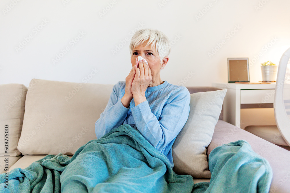 Mature woman with flu blowing nose at home. Close up elderly sick woman covered in blanket has cold blows her runny nose and suffers from flu condition. Mature woman with flu blowing nose at home. Close up elderly sick woman covered in blanket has cold blows her runny nose and suffers from flu condition.