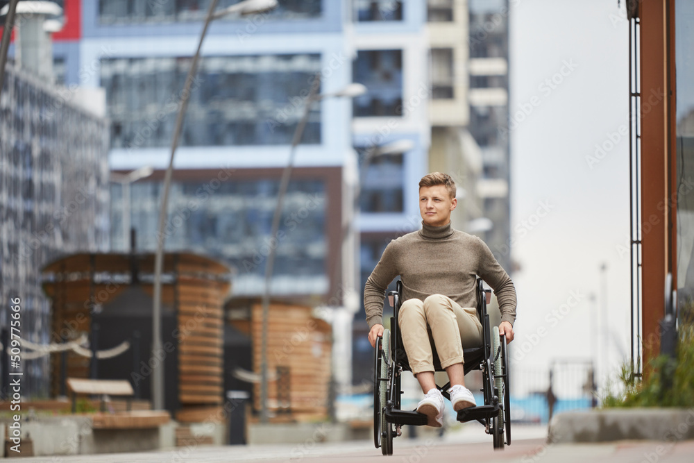 Content handsome young guy in casual clothing being mobile in wheelchair while moving over city street
