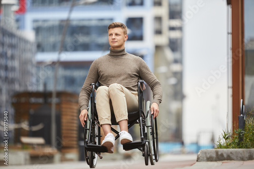 Content inspired young handicapped man sitting in wheelchair and crossing city district while walking alone