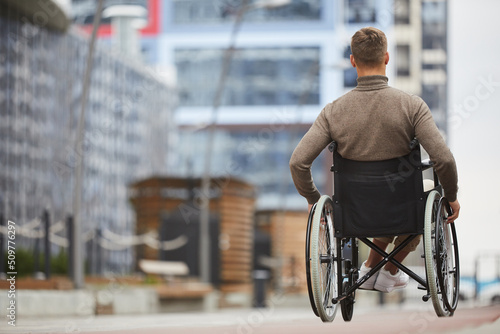 Rear view of young man in brown turtleneck pushing hand rims of wheelchair while riding wheelchair in city