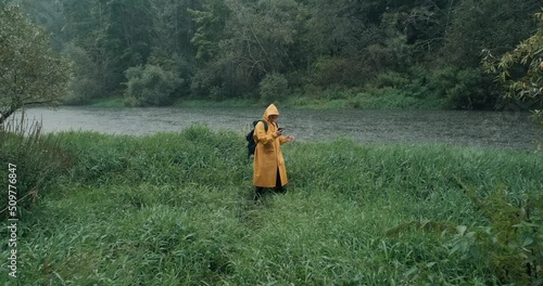 Heavy rain, hiker in yellow raincoat with backpack in tall grass next to river. The man has a phone with navigator in his hands. The concept of backpacking, lifestyle, hike, extreme hobbies and travel