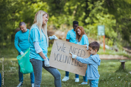Young volunteers collect garbage in nature. They hold a “We need volunteers” sign pointing at the camera.
