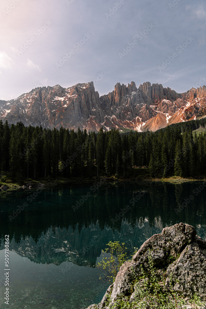 Vertical photo of a lake in the middle of the nature in the Dolomites, surrounded by trees, hill, mountains and snow. The water of the lake is clean and transparent. Refrection of the mountain.