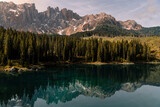 Horizontal photo of a lake in the middle of the nature in the Dolomites, surrounded by trees, hill, mountains and snow. The water of the lake is clean and transparent. Refrection of the mountain.