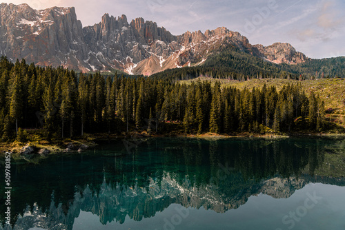 Horizontal photo of a lake in the middle of the nature in the Dolomites, surrounded by trees, hill, mountains and snow. The water of the lake is clean and transparent. Refrection of the mountain.