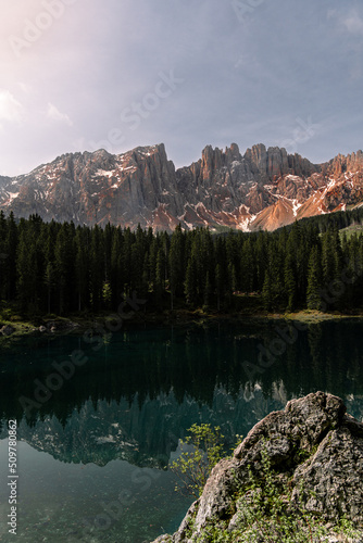 Vertical photo of a lake in the middle of the nature in the Dolomites, surrounded by trees, hill, mountains and snow. The water of the lake is clean and transparent. Refrection of the mountain.