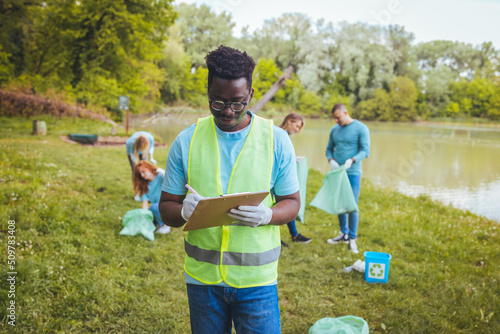 Front view of a smiling young man holding a clipboard in a vest. Happy male volunteer cleans the park. He is a volunteer and helps maintain the park