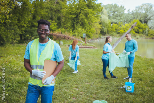 Portrait of a smiling young man holding a clipboard in a vest. Happy male volunteer cleans the park. He is a volunteer and helps maintain the park