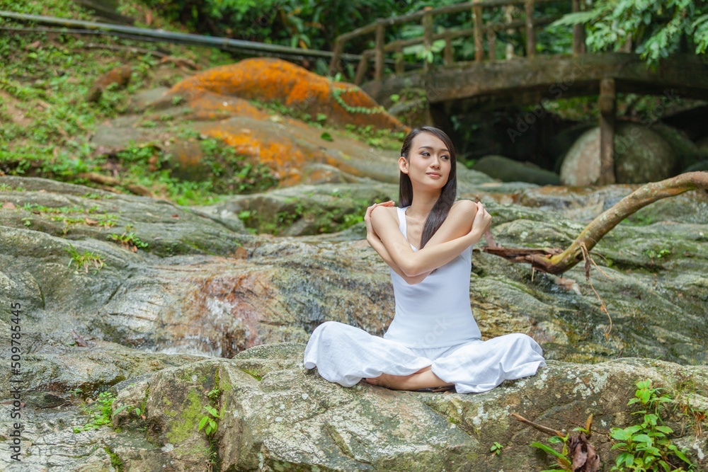 Asian young female lady yoga model meditate and practicing yoga zen poses in a nice, quiet tranquil green outdoor forest park with giant stone, tree and nice little stream waterfall Asian young female lady yoga model meditate and practicing yoga zen poses in a nice, quiet tranquil green outdoor forest park with giant stone, tree and nice little stream waterfall