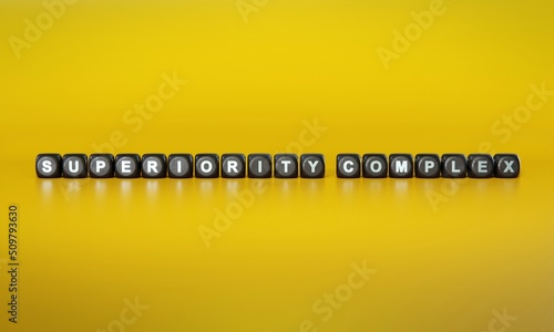 Words ‘superiority complex’ spelled out in white text on dark wooden blocks against plain yellow background. 3D rendering © HTGanzo