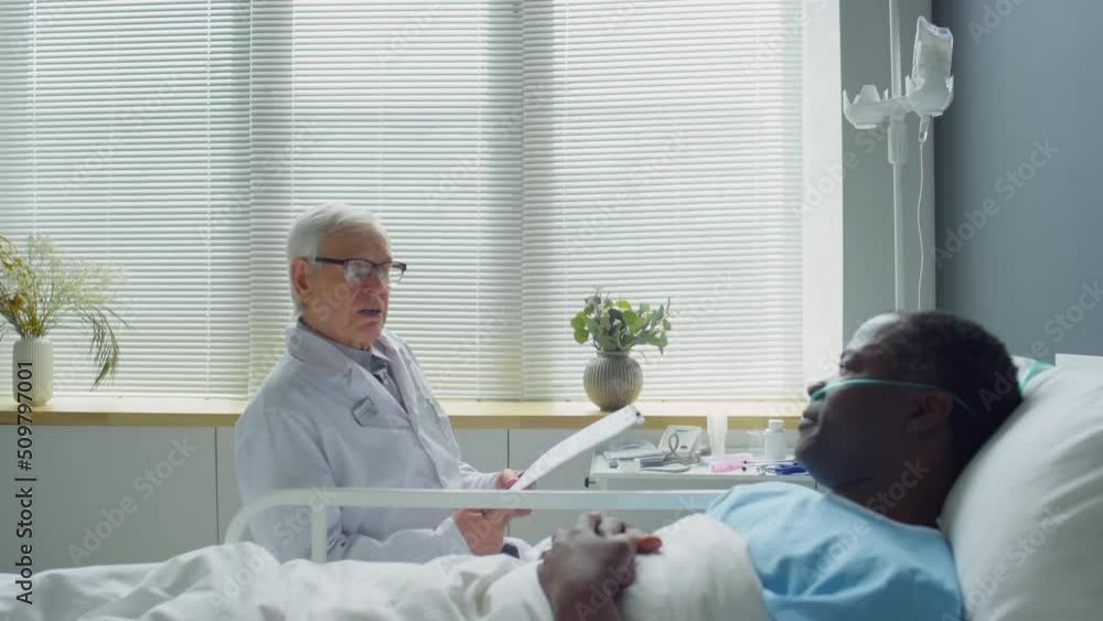 Zoom out shot of senior Caucasian doctor walking in hospital ward, sitting at bedside and talking with African American patient lying with nasal cannula Zoom out shot of senior Caucasian doctor walking in hospital ward, sitting at bedside and talking with African American patient lying with nasal cannula