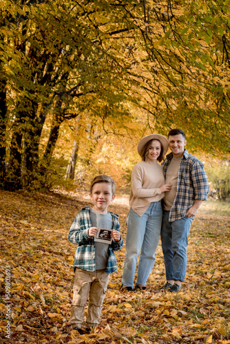 Happy family of a man with a woman and surprised son holding an ultrasound pregnancy picture. Autumn photo shoot of a family waiting to be replenished