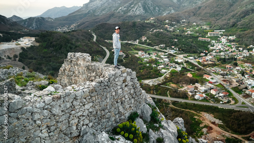 A young guy stands on the edge of the ruined Haj Nehai fortress against the backdrop of a valley and mountains. The guy poses against the backdrop of the mountains of Montenegro. Fortress in Sutomore