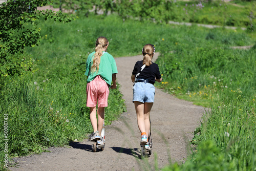 Two kid girls on electric scooters go downhill in summer park